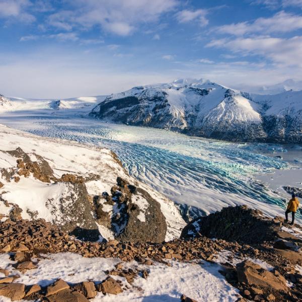 Person in a yellow jacket overlooking a vast glacial valley with blue ice and snowy mountains under a clear sky.