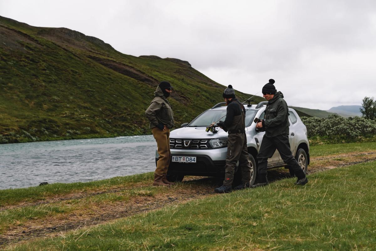 Dacia Duster parked on grass beside a river with three people gathered at the front