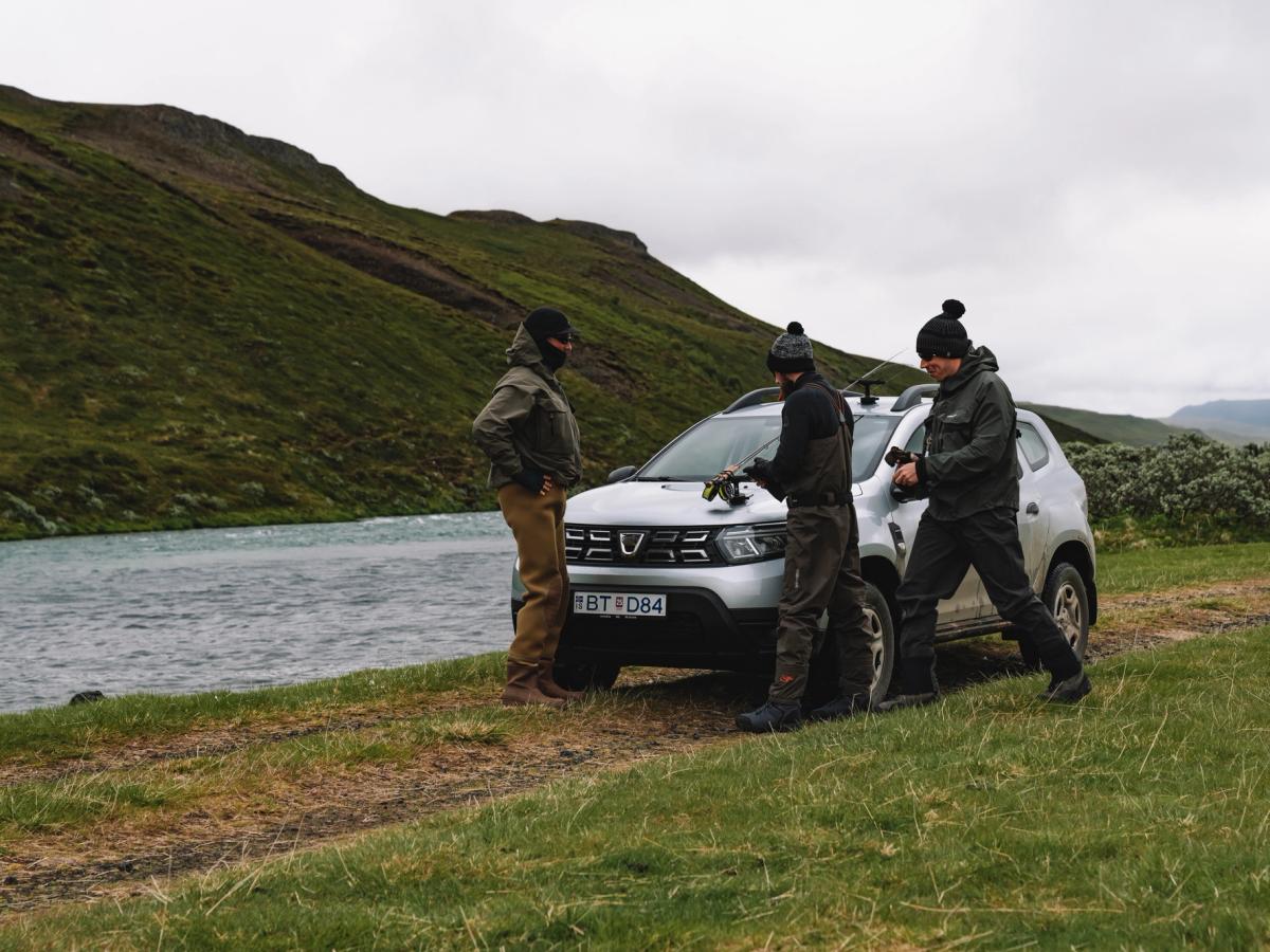 Grupo Dacia Duster Riverside Dacia Duster aparcado sobre la hierba junto a un río, con tres personas reunidas delante