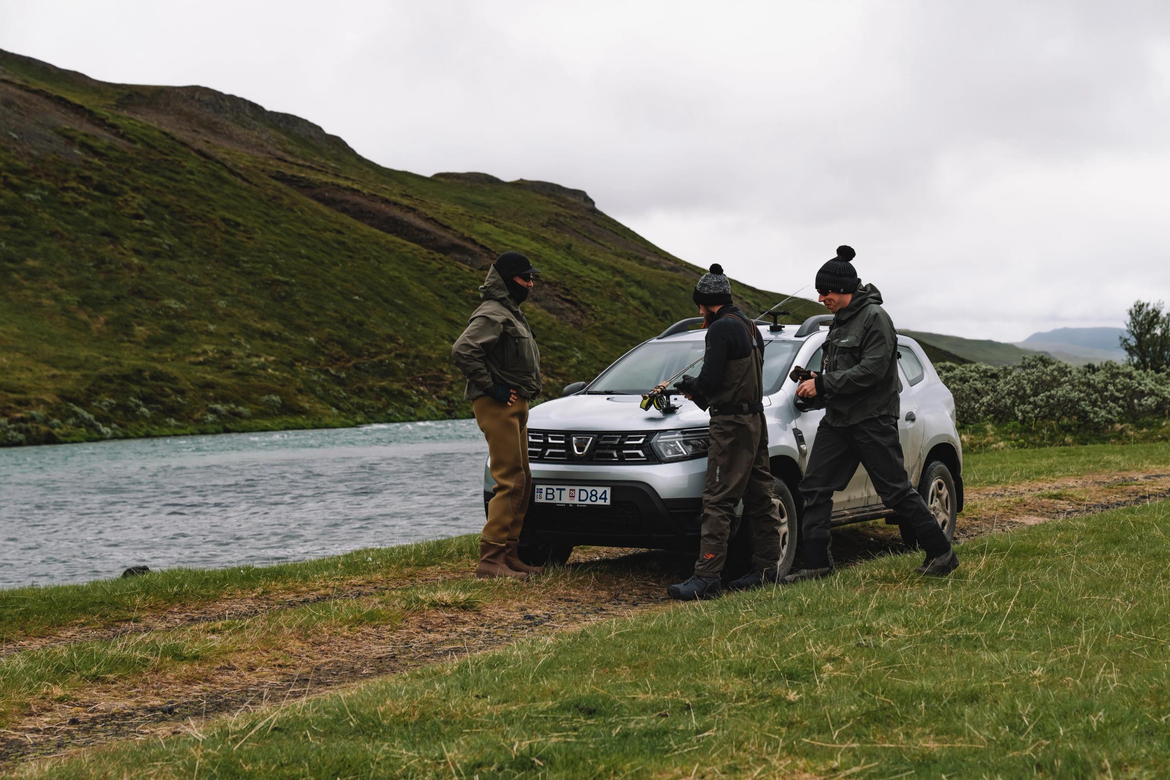 Grupo Dacia Duster Riverside Dacia Duster aparcado sobre la hierba junto a un río, con tres personas reunidas delante