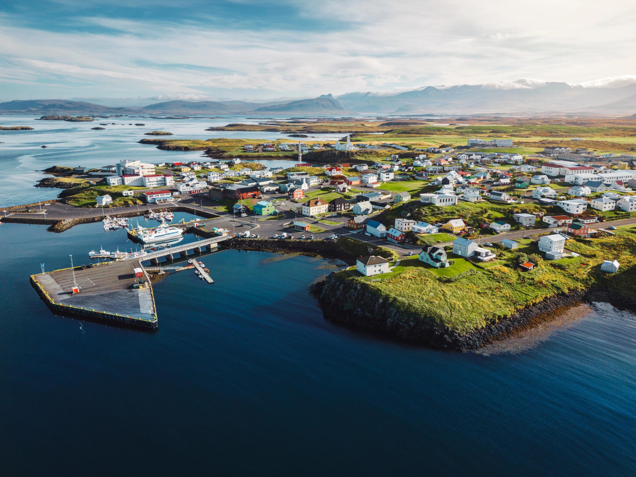 Aerial view of a colorful coastal town with a harbor, boats, green islands, and distant mountains.