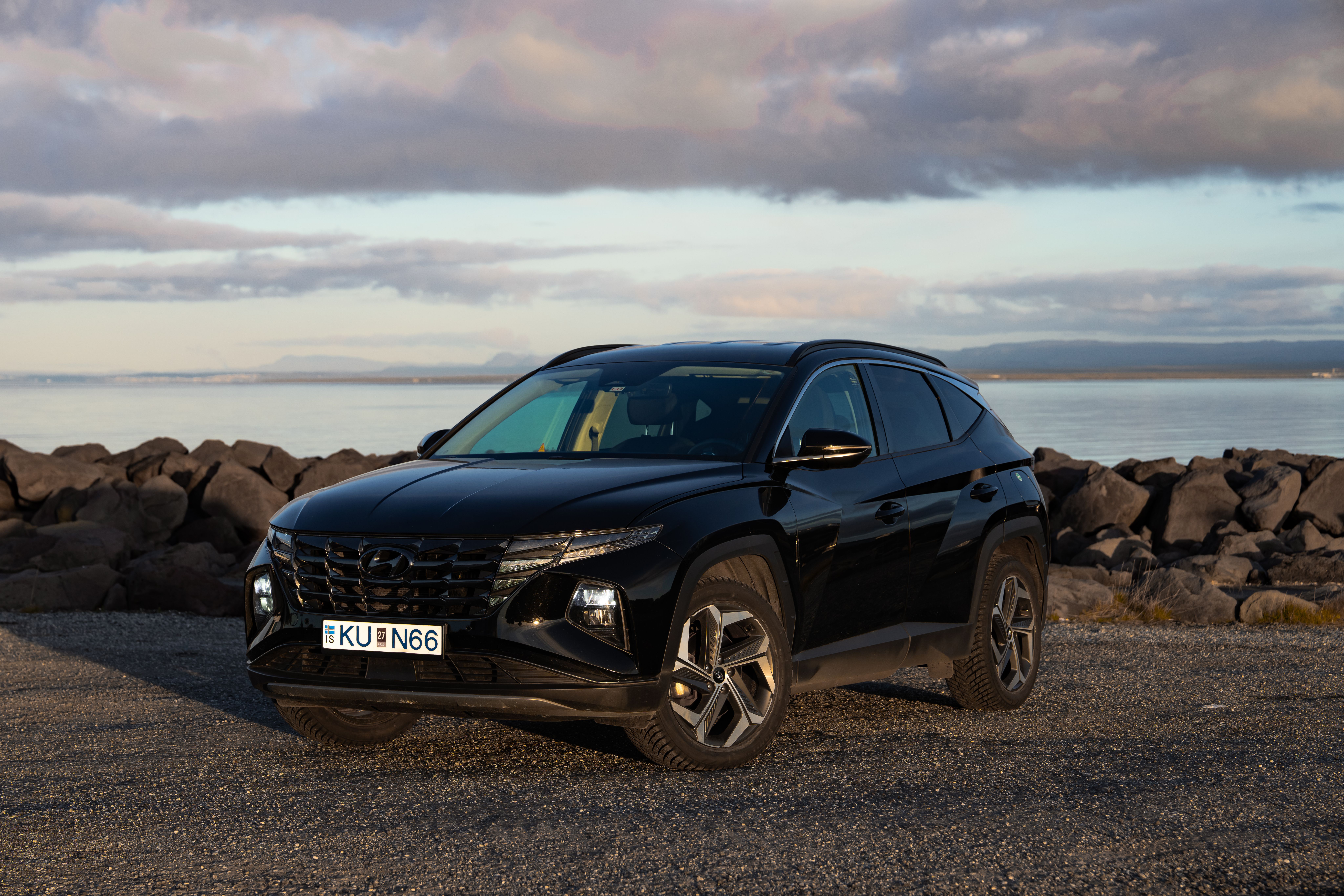 Hyundai Tucson noir garé sur du gravier au bord d’une côte rocheuse, avec des montagnes de l’autre côté de l’eau