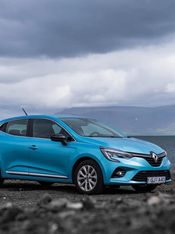 A turquoise Renault Clio car parked on a rocky shore by a lake with mountains under a cloudy sky.