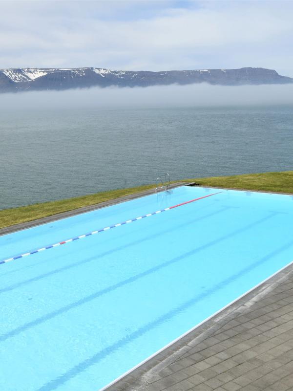 A bright blue outdoor swimming pool overlooking a misty ocean and snow-capped mountains.