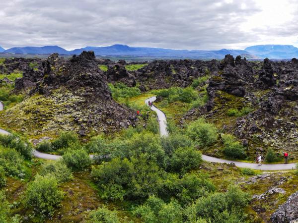 A panoramic view of Iceland's Dimmuborgir lava field, with unique black rock formations, vibrant green vegetation, winding paths, and distant mountains.