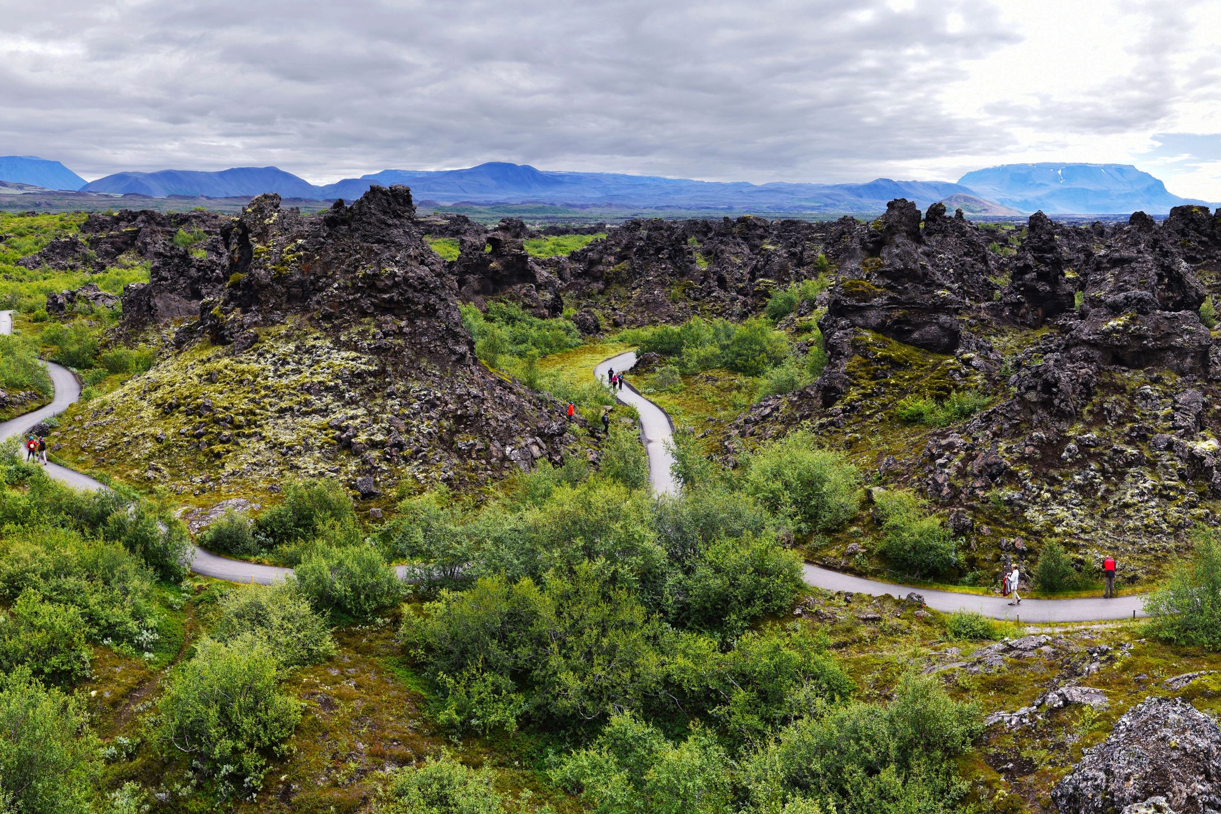 A panoramic view of a volcanic landscape with dark, jagged rock formations, green moss and shrubs, and winding paths with people.