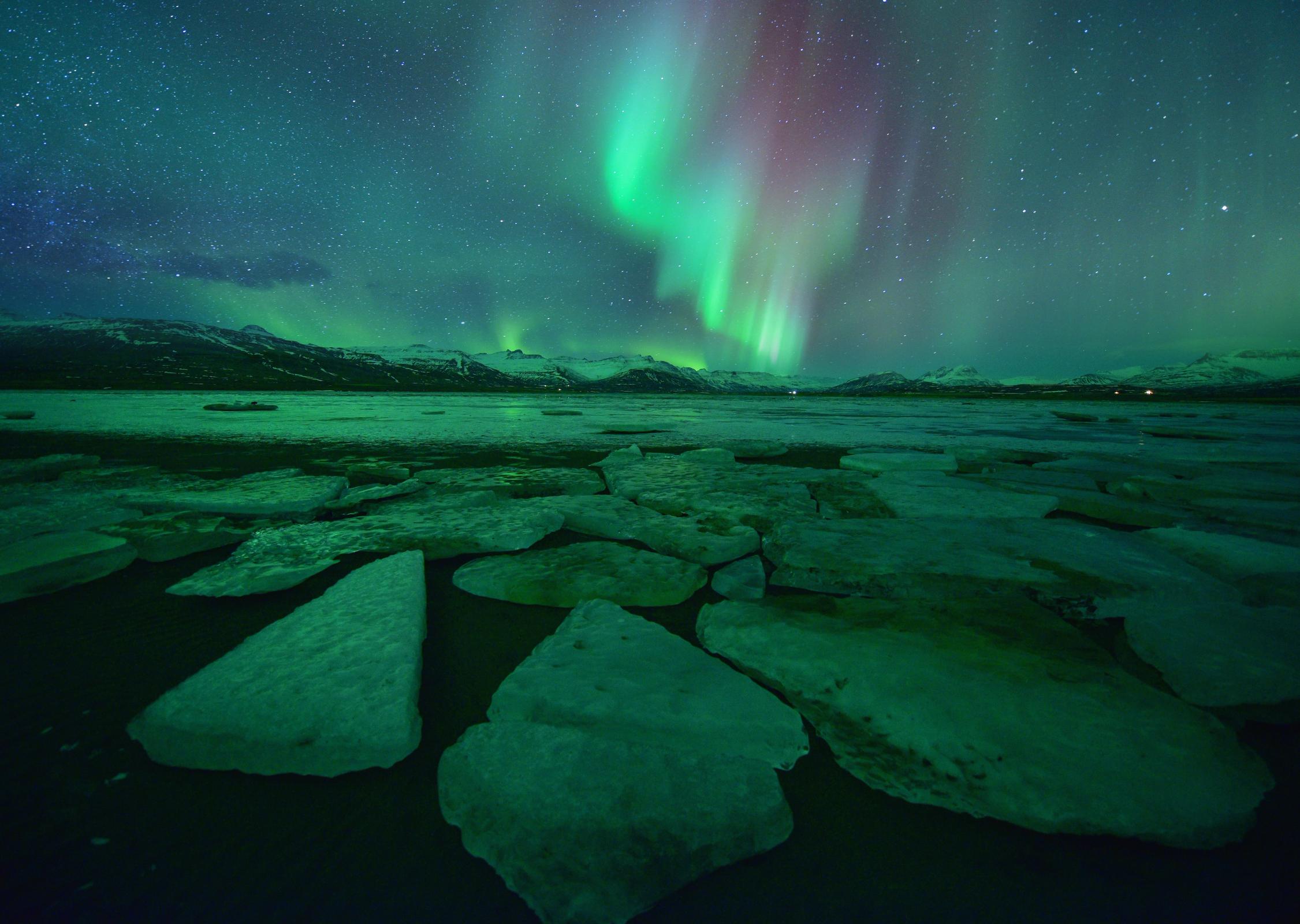 Aurora boreal, verde y roja, sobre un cielo estrellado, montañas nevadas, un lago oscuro y grandes bloques de hielo en primer plano.