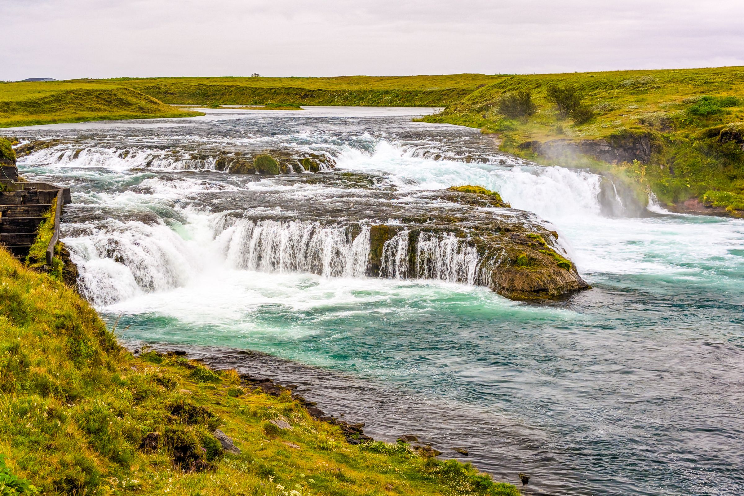 Cascada de varios niveles con agua turquesa fluyendo a través de un paisaje verde y herboso bajo un cielo nublado.