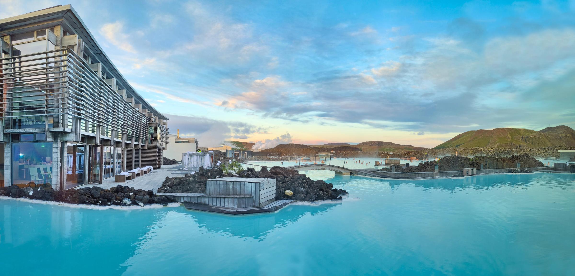 Panoramic view of a modern building beside a milky blue geothermal lagoon with steam rising from the water and dark volcanic rocks.