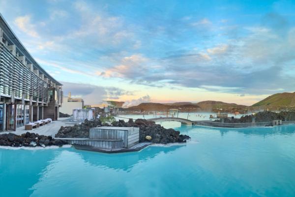 A modern building stands next to a wide, milky blue geothermal pool with steam rising, surrounded by dark volcanic rocks under a cloudy sky.