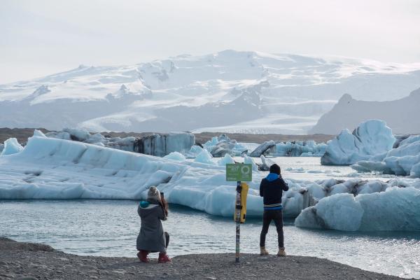 Two people photograph icebergs in a glacial lagoon with snowy mountains in the background.