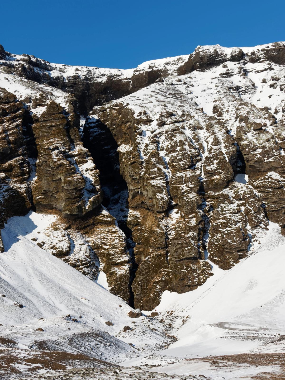 A snow-covered rocky mountain with a deep fissure, and a person standing at its base under a clear blue sky.