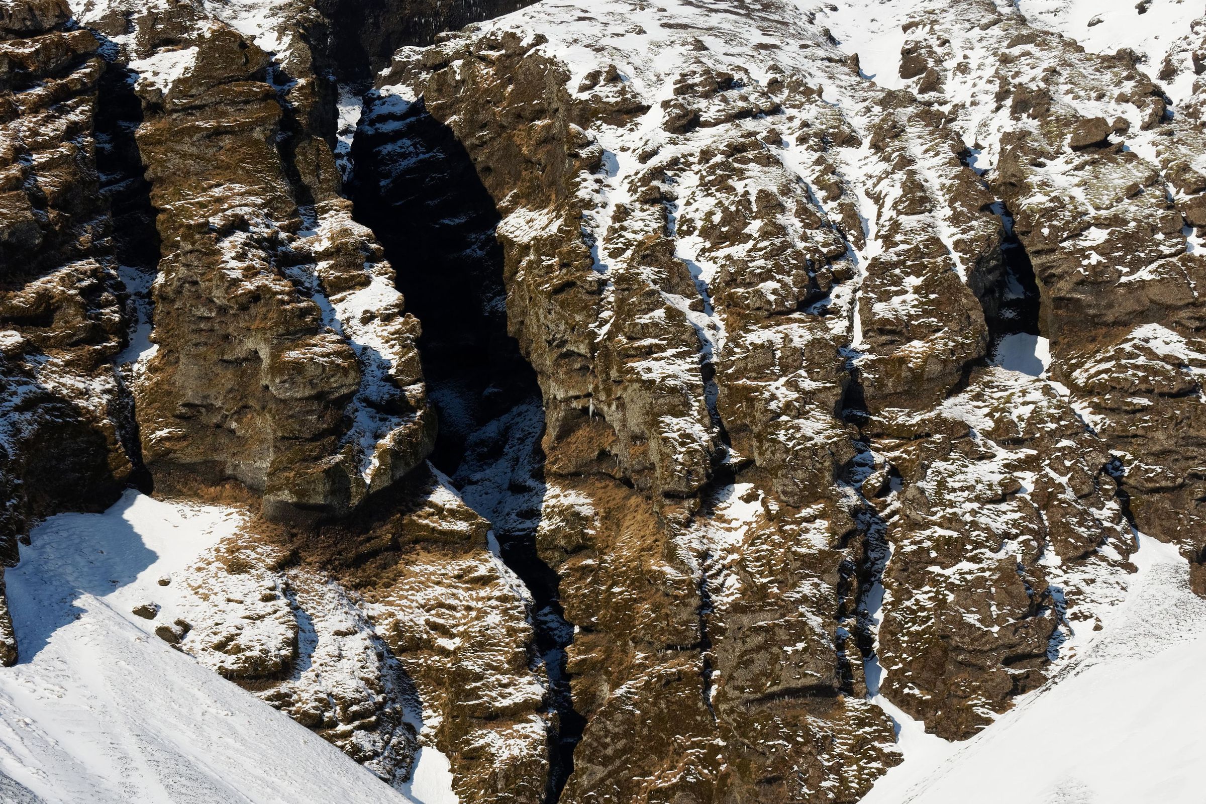 A snow-covered rocky mountain with a deep fissure, and a person standing at its base under a clear blue sky.