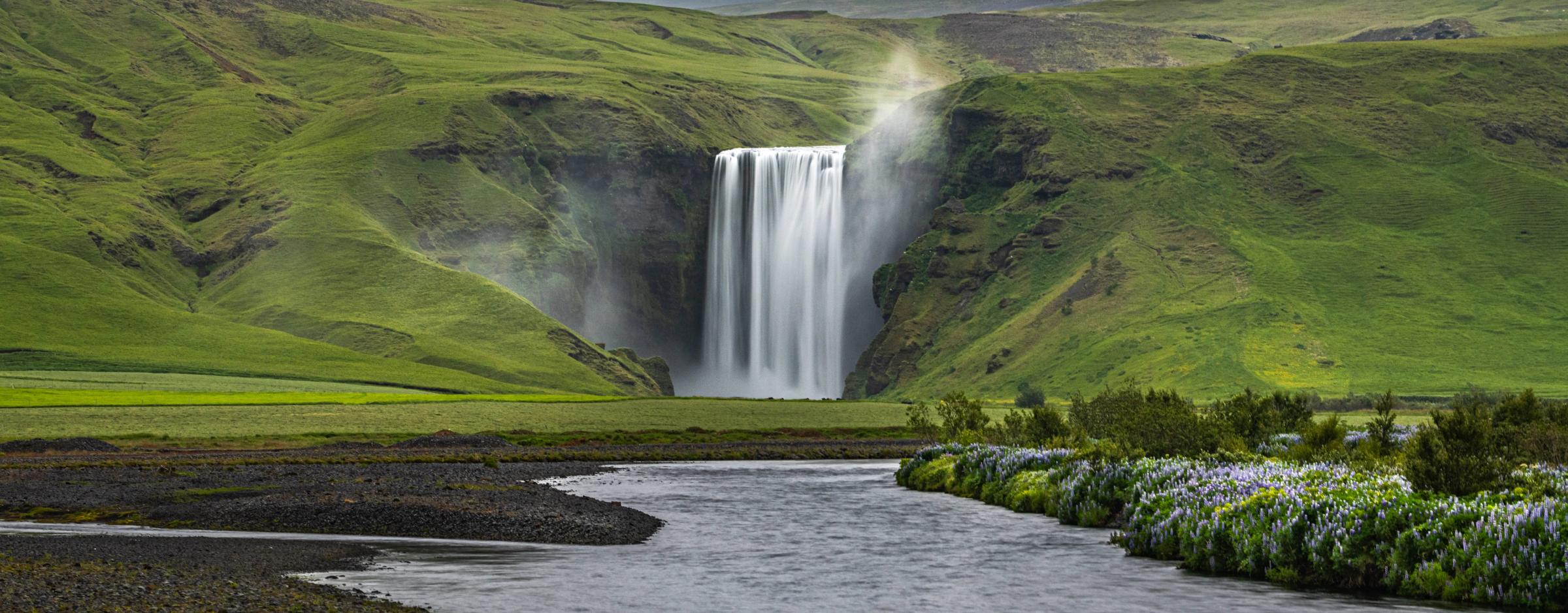 una cascada rodeada de colinas verdes, con un río delante y flores silvestres en su margen