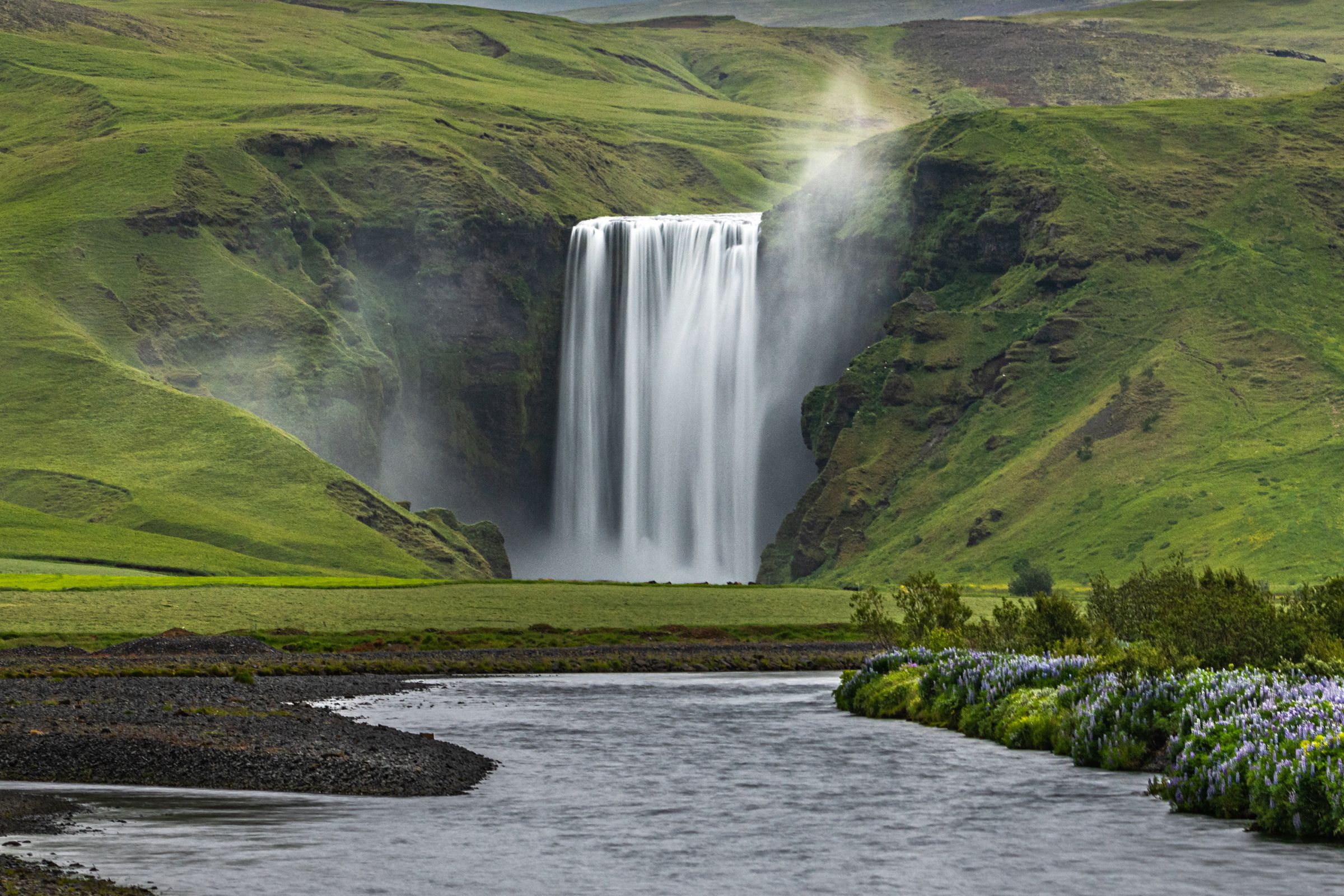 Una cascada se precipita en un río, rodeada de verdes colinas y orillas florecientes.