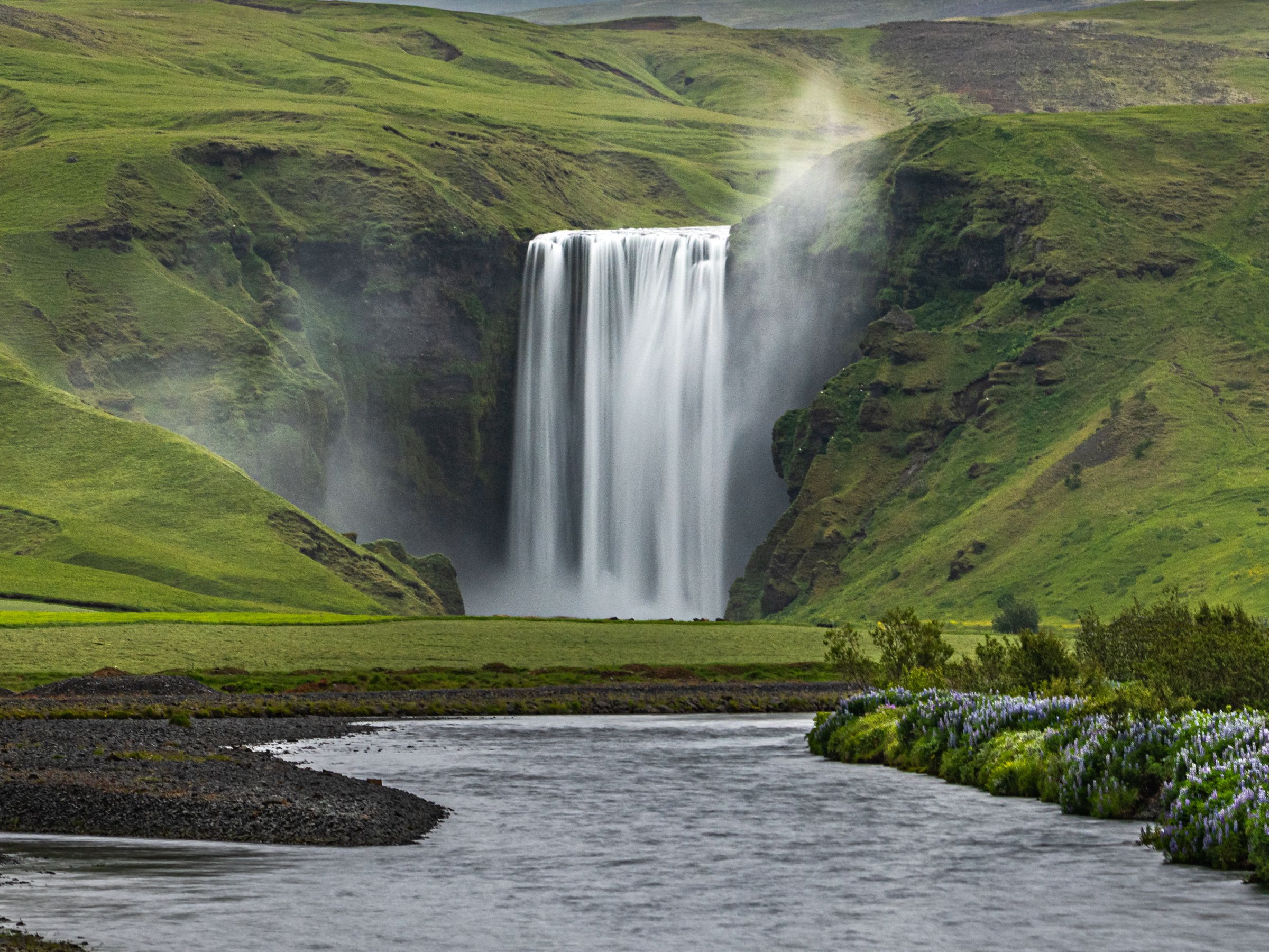 una cascada rodeada de colinas verdes, con un río delante y flores silvestres en su margen