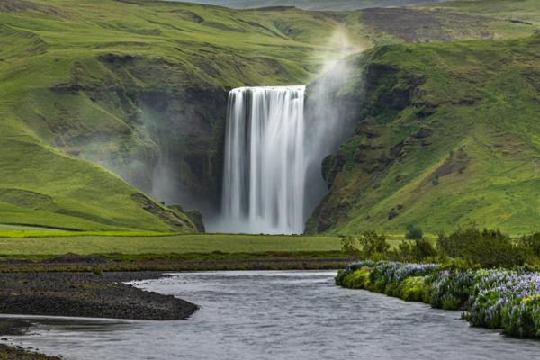 panoramic of a powerful waterfall surrounded by moss covered mountains and a river on the foreground