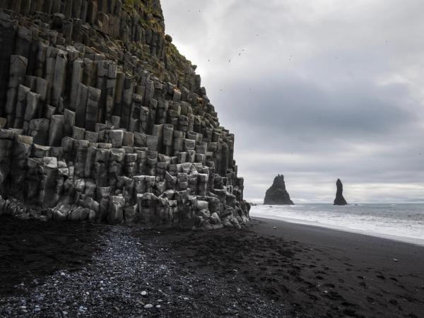 Black sand beach with basalt columns and two sea stacks under a cloudy sky.