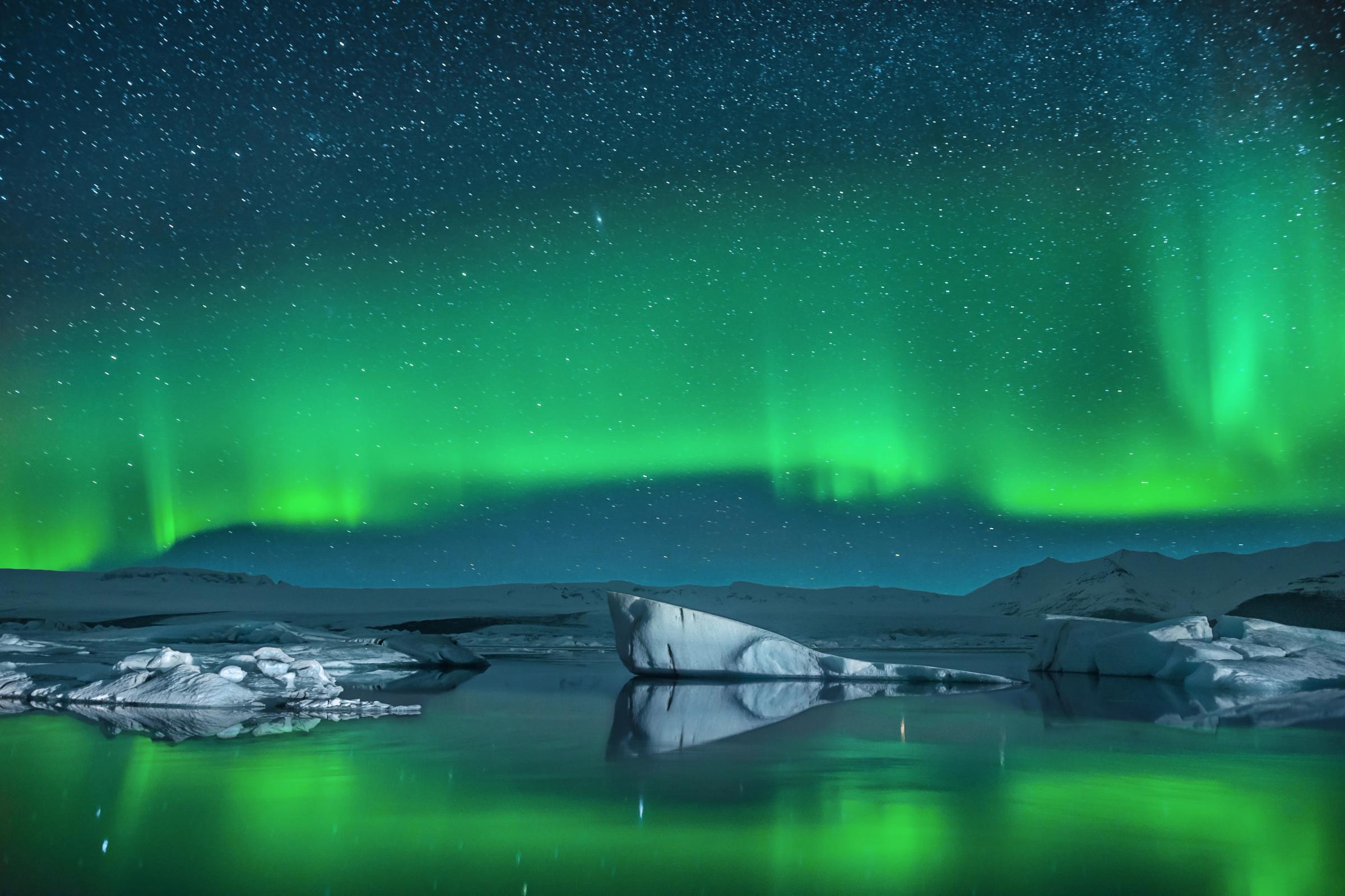 Vibrant green aurora borealis illuminates a starry sky, reflecting in a lake with icebergs.