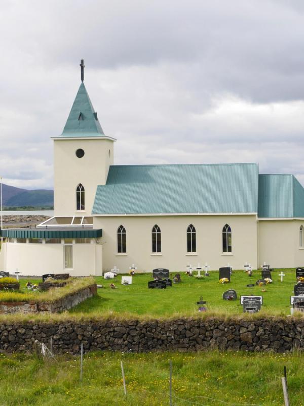 A cream-colored church with a teal roof and spire stands in a grassy cemetery, with a lake and dark mountain in the background under a cloudy sky.
