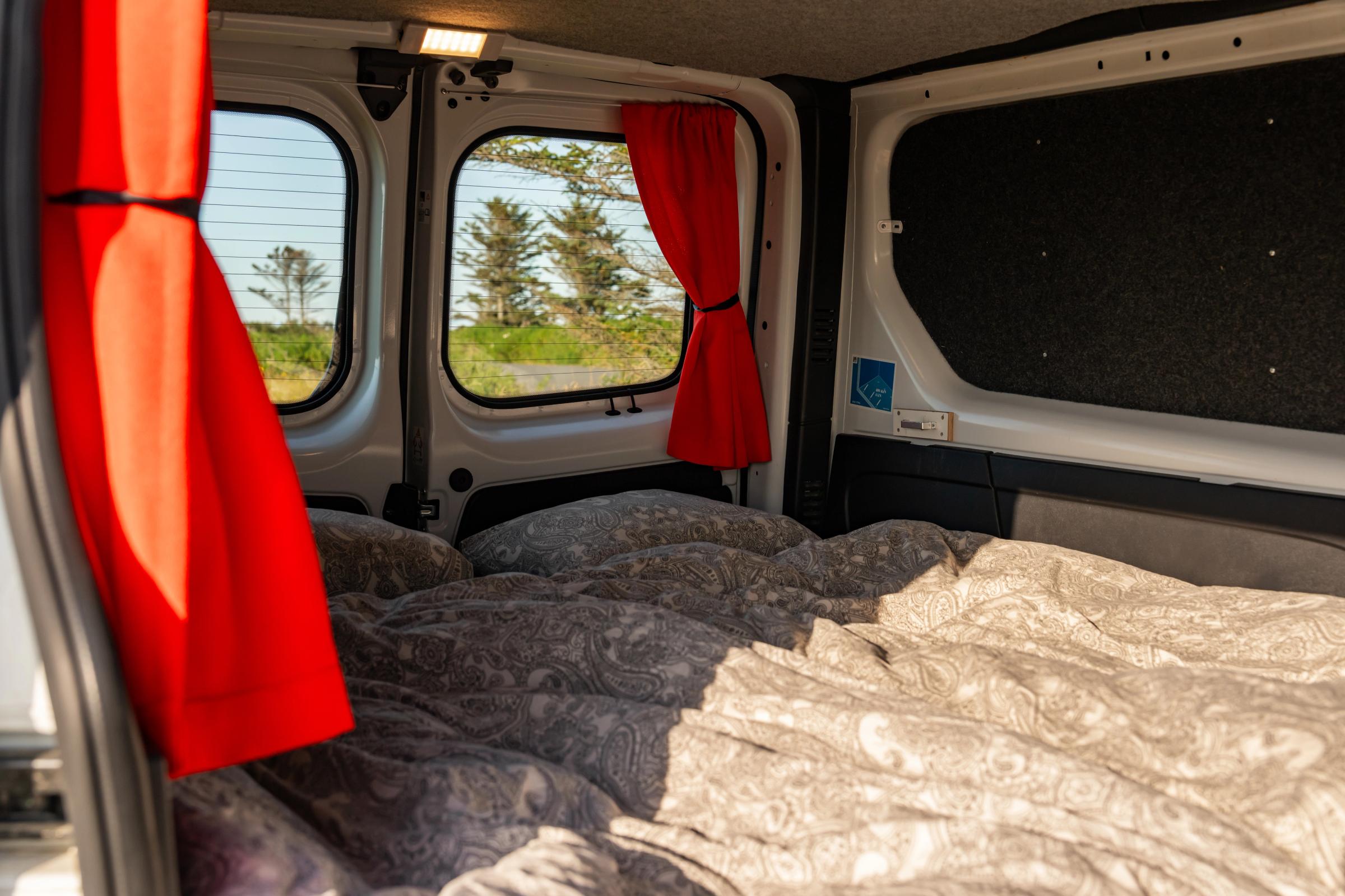 Bed with duvet and pillows facing the closed rear doors and their red curtained windows