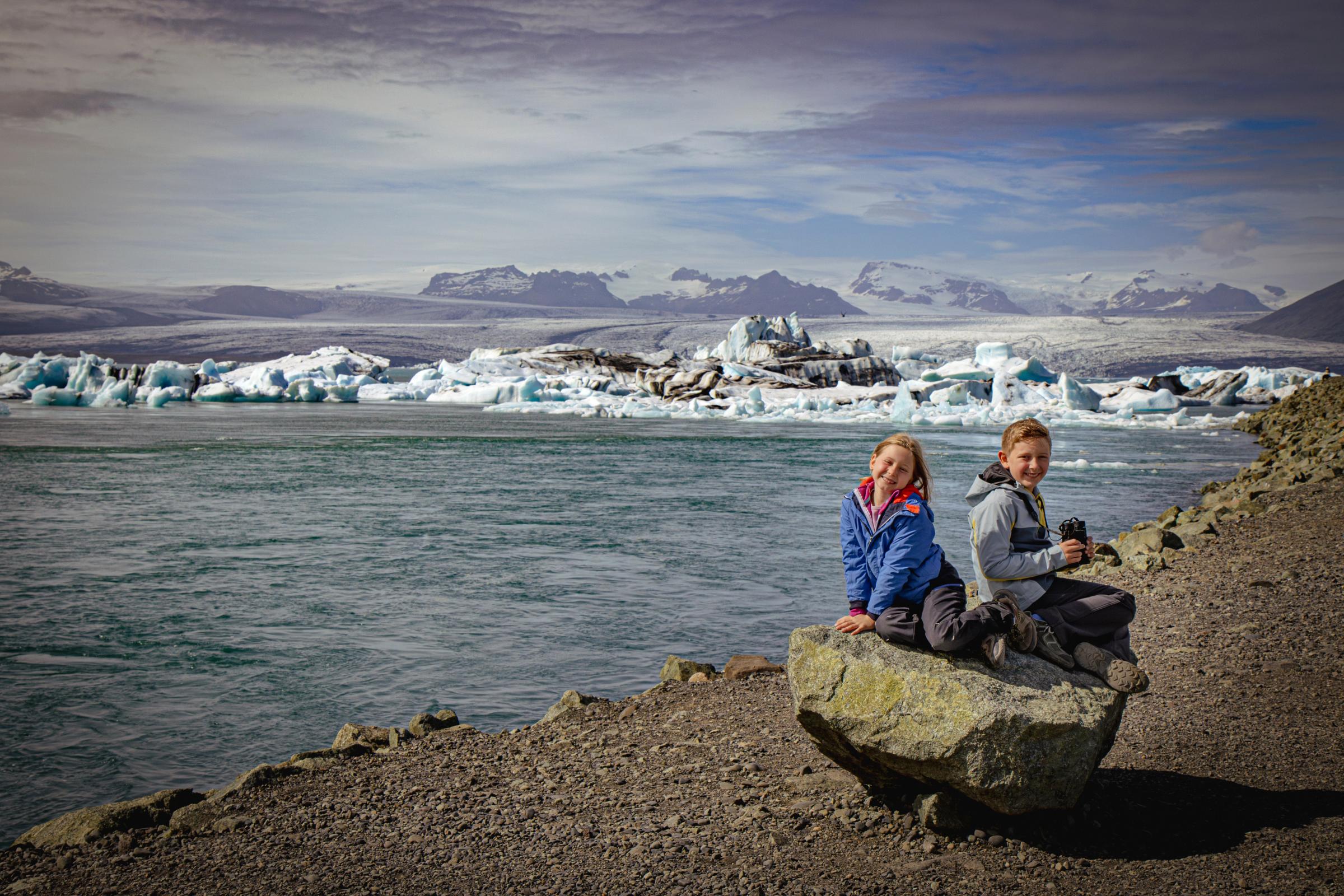Two smiling children sit on a rock by a glacial lagoon filled with icebergs, with mountains in the background.