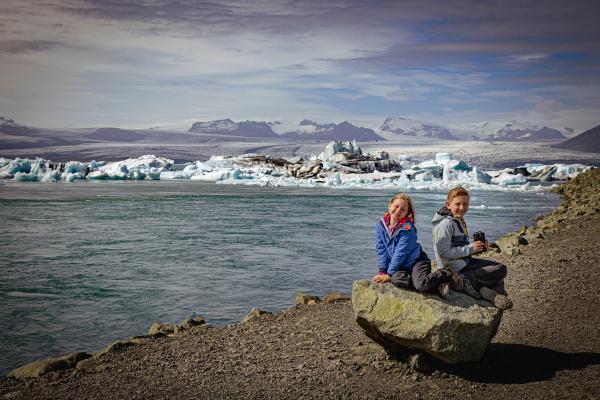 Two smiling children sit on a rock overlooking a glacial lagoon filled with icebergs and mountains.