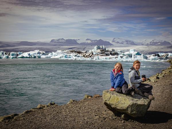 Two smiling children sit on a rock by a glacial lagoon filled with icebergs, with mountains in the background.