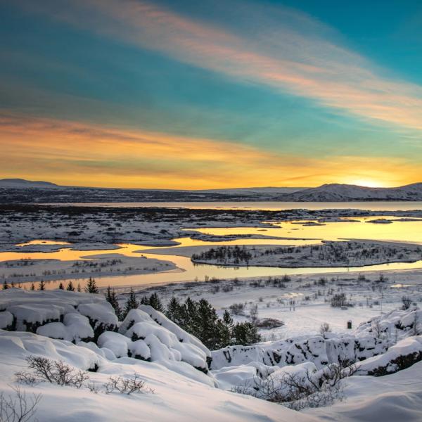 A vast snowy landscape with a winding lake reflecting a vibrant orange and blue sunset sky.