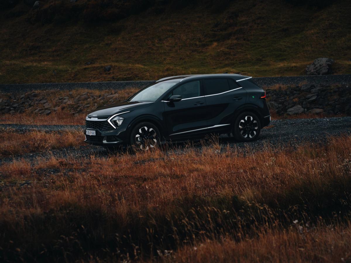 Vegaaðstoðarsnið Sportage Kia Sportage séður skáhallt að framan, lagt á malarkanti við veginn við hliðina á háu grasi