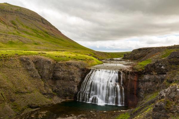 A wide waterfall cascades into a pool within a rocky canyon, flanked by green hills under a cloudy sky.