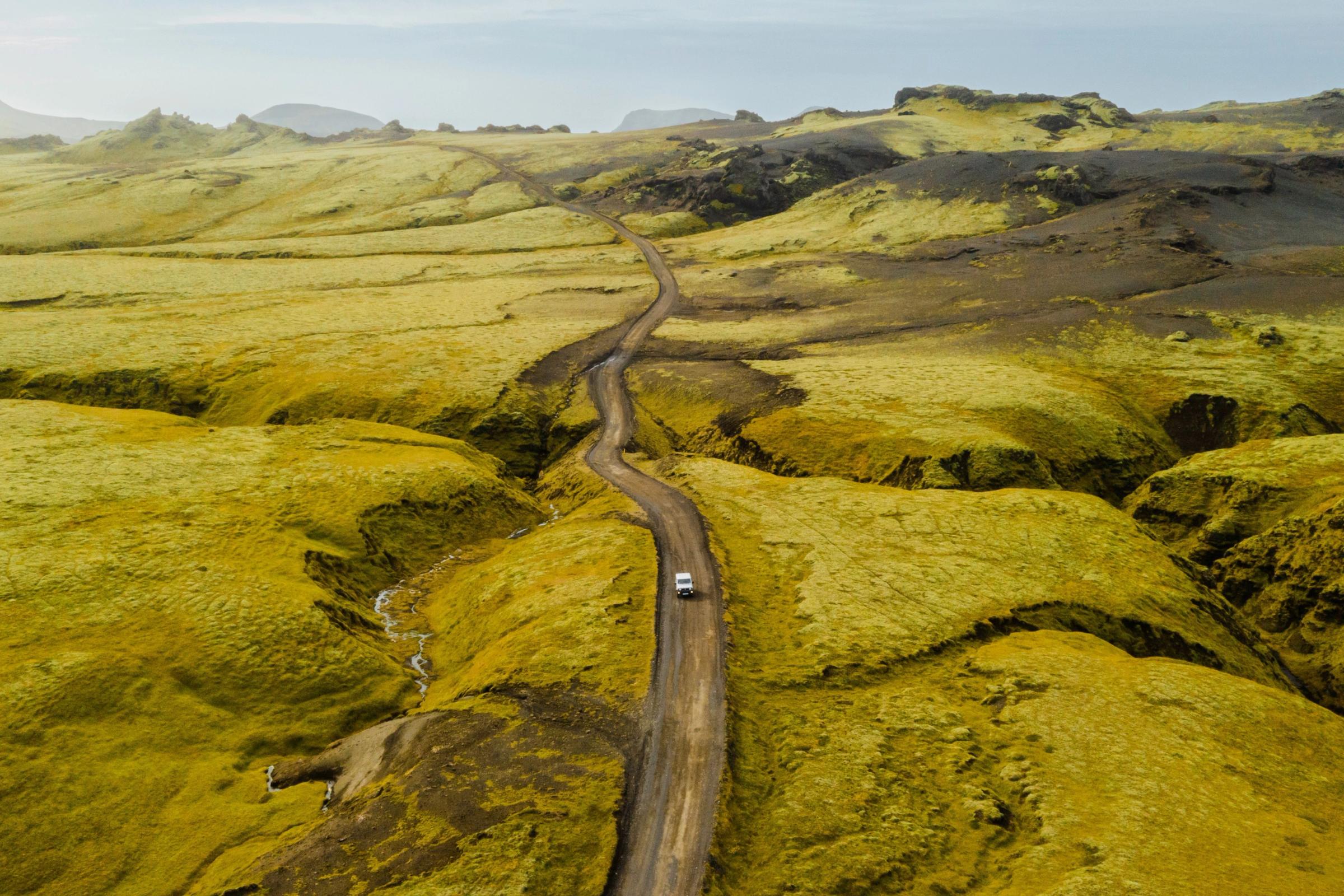 Vista aérea de un coche blanco en un camino de tierra serpenteante que atraviesa colinas verdes cubiertas de musgo.