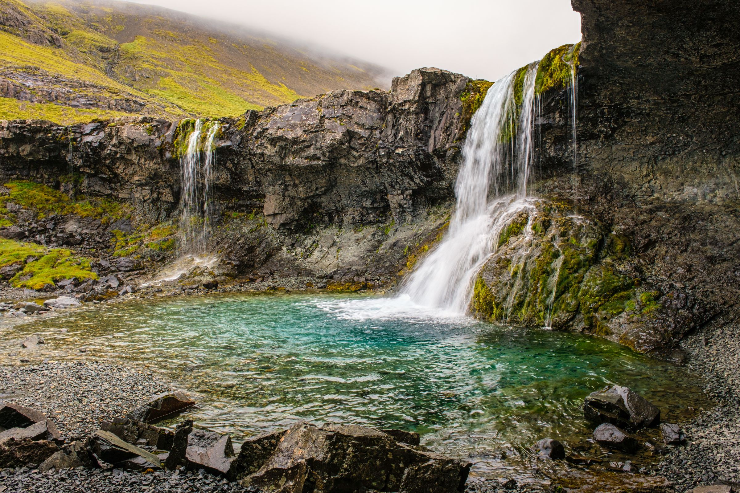 Two waterfalls cascade into a vibrant turquoise pool surrounded by mossy cliffs and a misty green mountain.