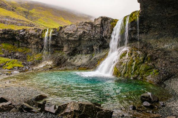 Two waterfalls cascade into a vibrant turquoise pool surrounded by mossy cliffs and a misty green mountain.