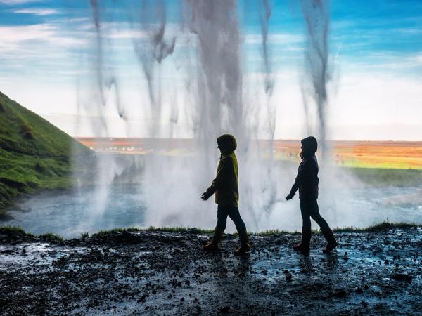 Two silhouetted people walk on a wet path behind a large waterfall, with a green hill and distant fields visible through the mist.