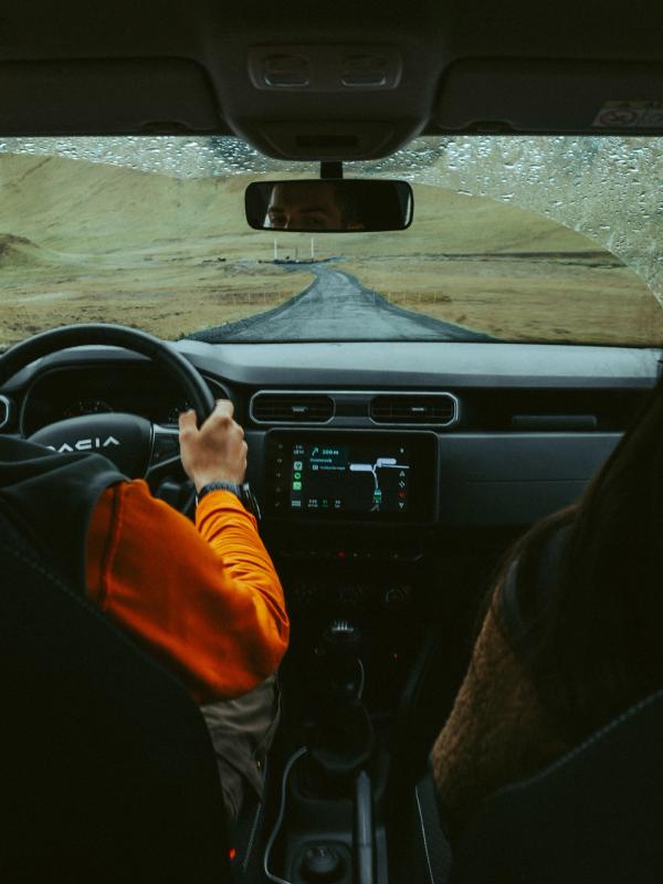 A person drives a car on a winding road through a hilly landscape, seen from the back seats with raindrops on the windshield.