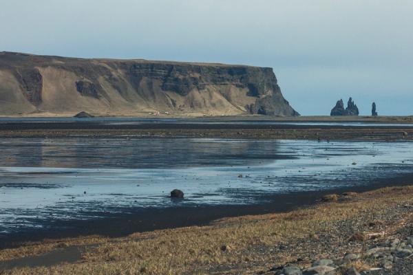 Coastal landscape with dark wet ground, a large cliff, and three prominent sea stacks in the distance.