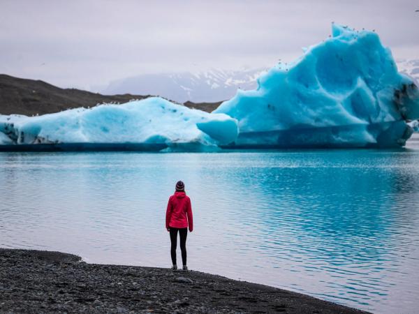 Mujer con una chaqueta roja en orilla de una laguna viendo un iceberg