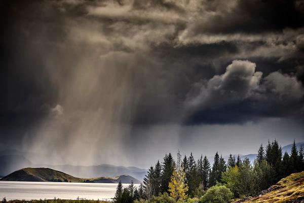 a storm is coming over a lake with trees in the foreground and mountains in the background .