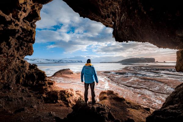 A person in a blue jacket stands at a cave entrance, looking out at a vast winter landscape with mountains and water.