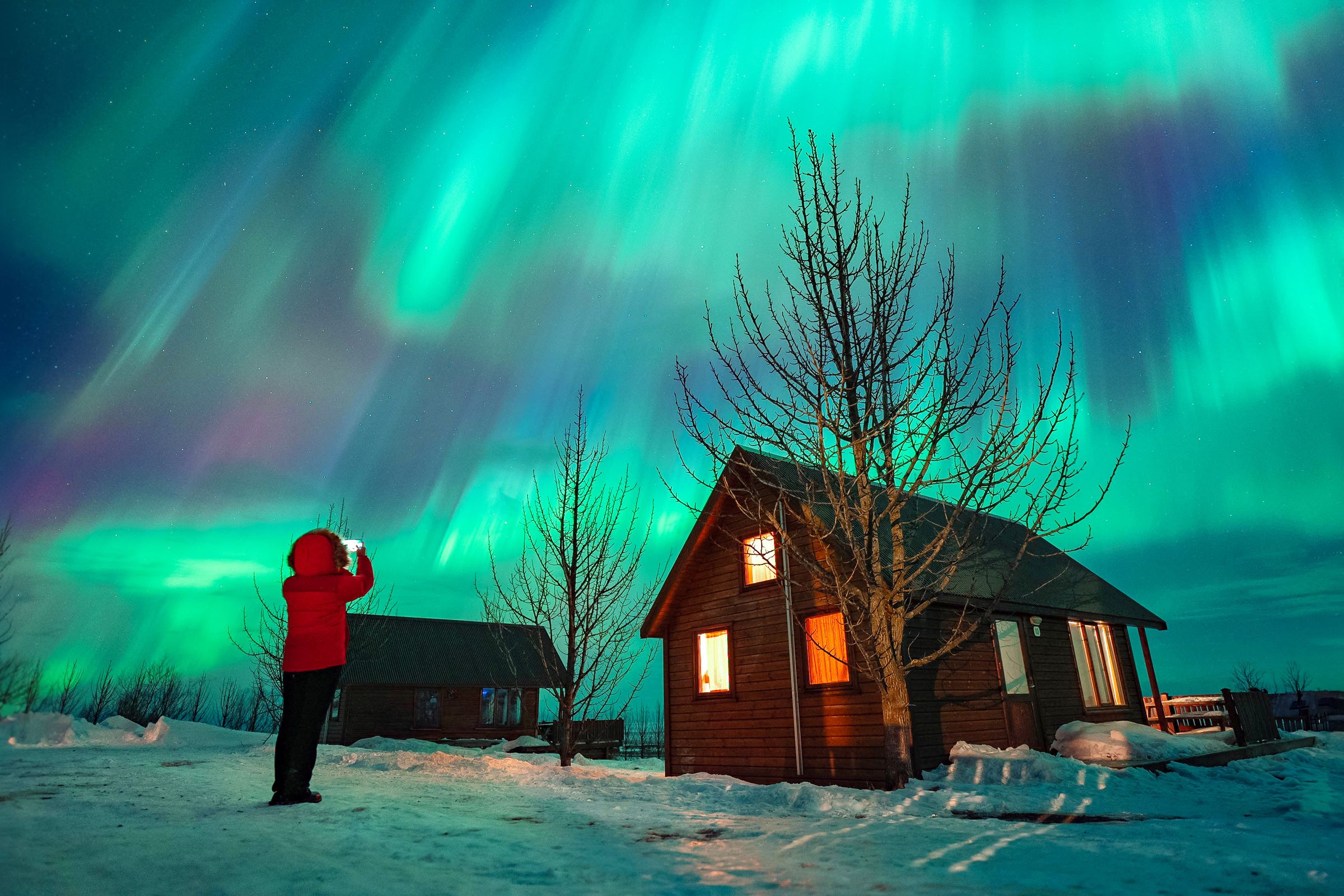 Una persona fotografía unas vibrantes auroras boreales verdes sobre unas cabañas cubiertas de nieve.