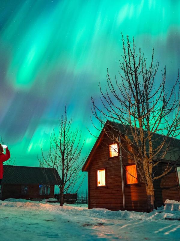 Person in a red jacket photographs vibrant green Northern Lights above snowy cabins.