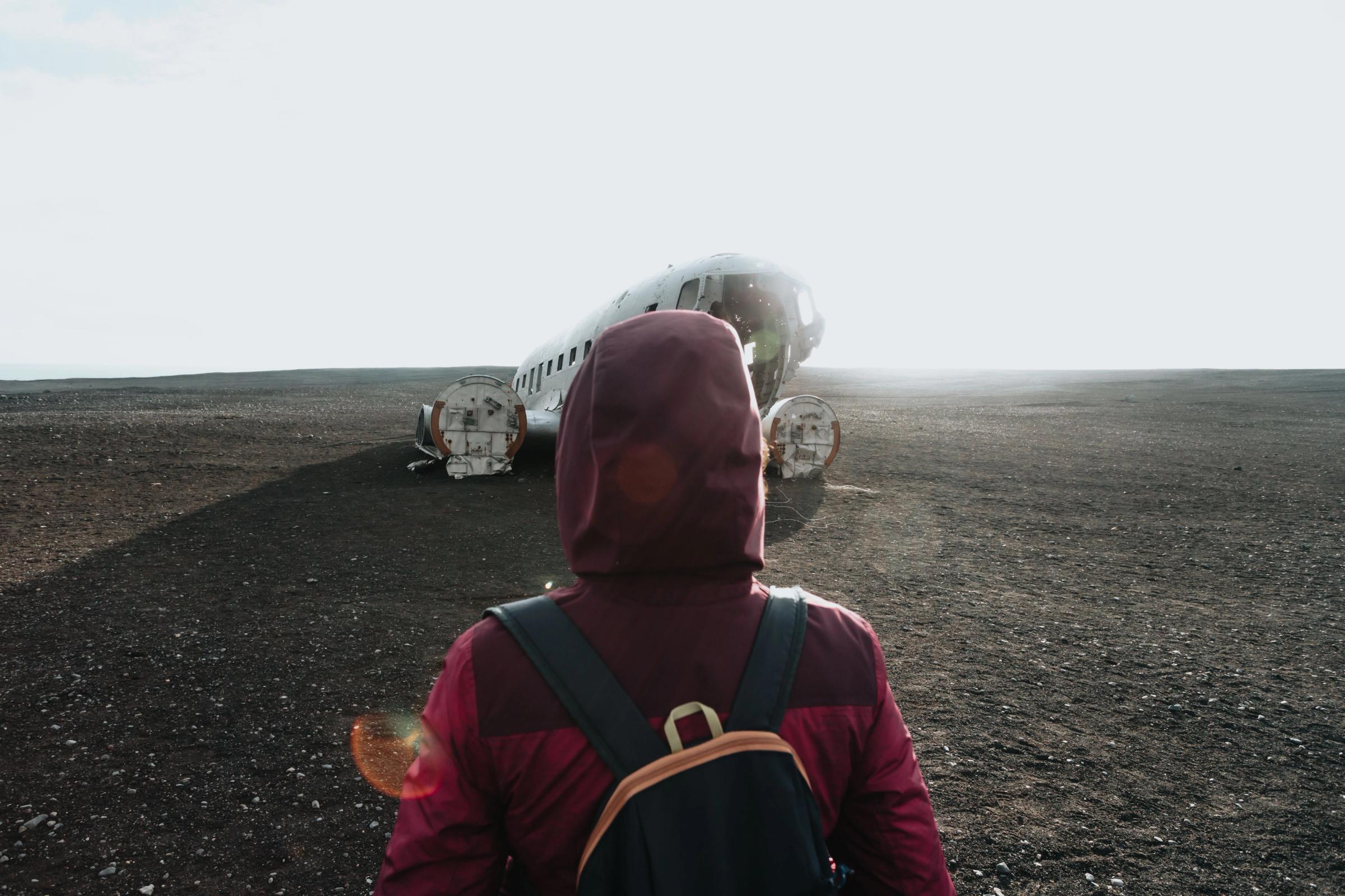 A person with a backpack walks toward a crashed airplane fuselage on a desolate black landscape.