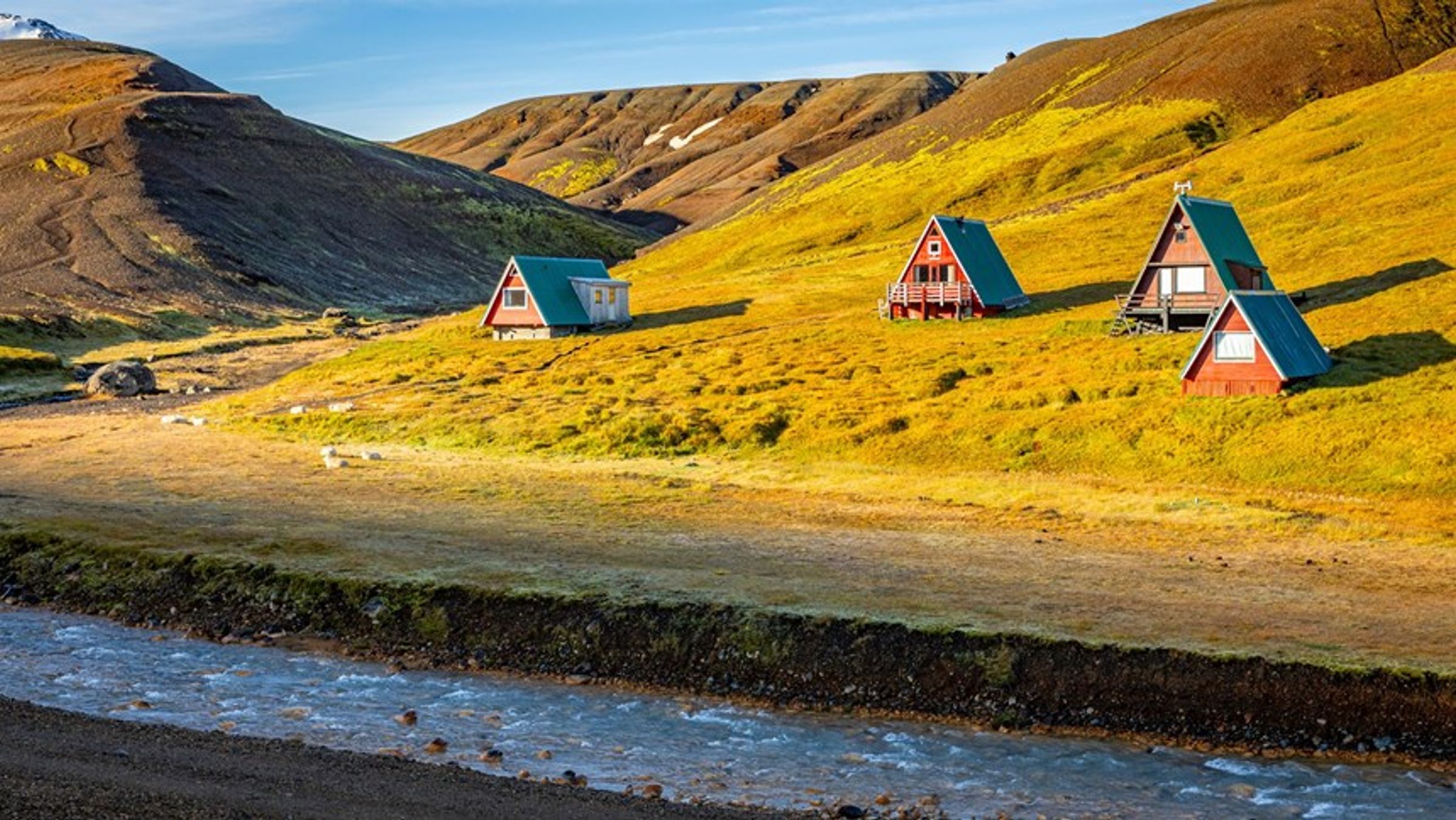 three small houses are sitting on top of a grassy hill next to a river at kerlingarfjöll campsite in iceland.