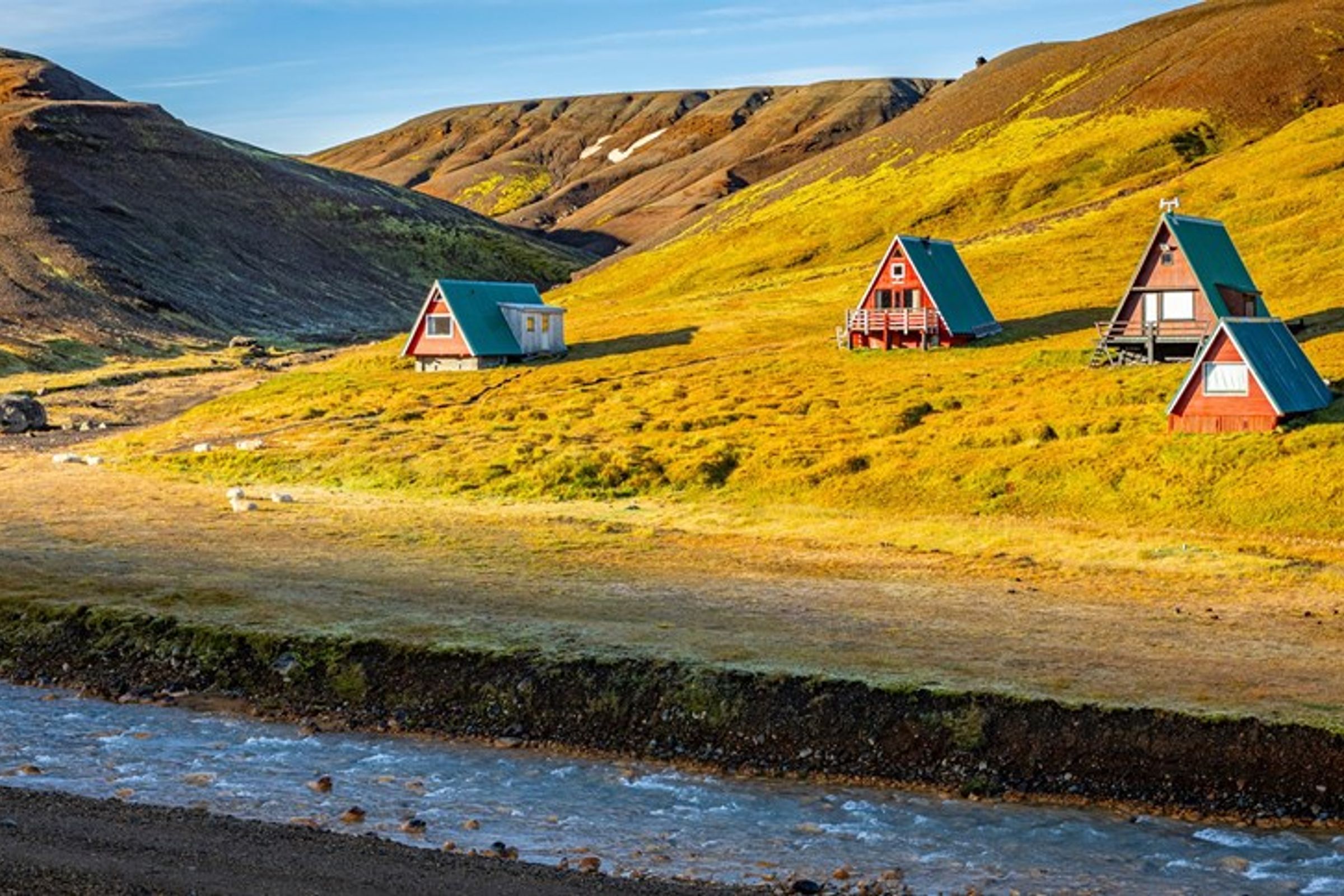 three small houses are sitting on top of a grassy hill next to a river at kerlingarfjöll campsite in iceland.