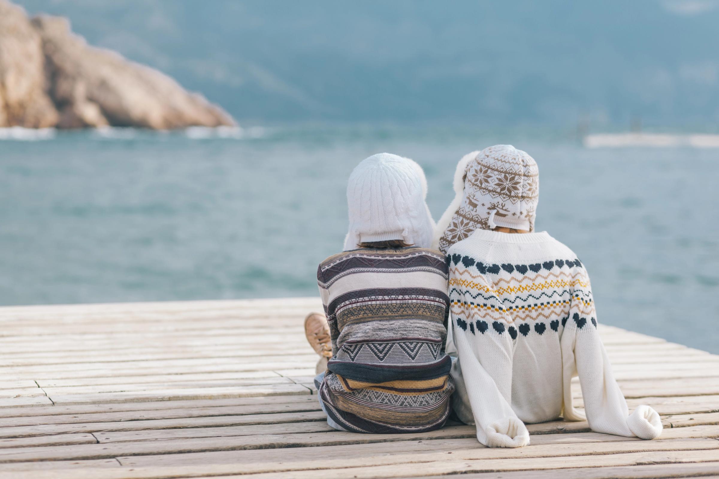 Dos personas con suéteres cálidos y gorros están sentadas en un muelle de madera, contemplando el agua y las montañas a lo lejos.