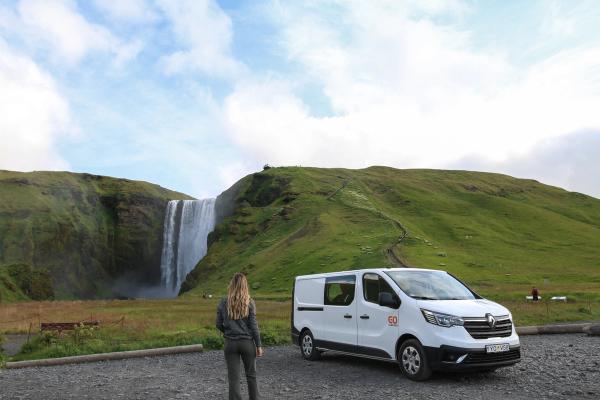 A woman faces a large waterfall cascading down green hills, with a white rental van parked in a gravel lot beside her.