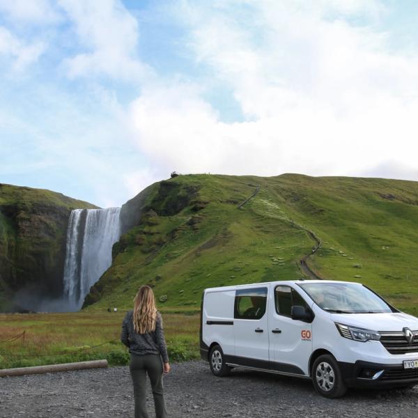 A person stands in a gravel lot, looking at a large waterfall flanked by green hills, with a white camper van parked nearby.