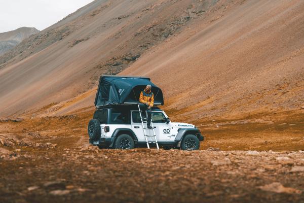 A person sits on an open rooftop tent on a white Jeep Wrangler in a barren, mountainous landscape.