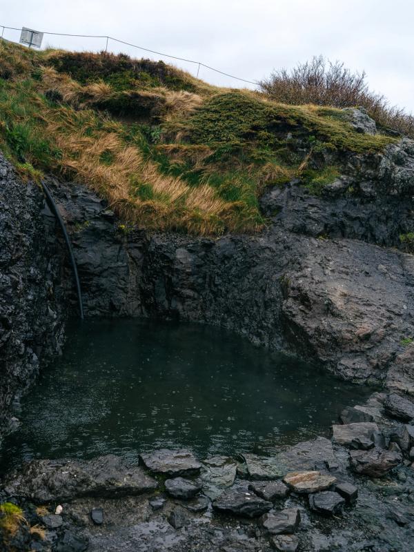 A natural pool among dark rocks and a grassy hill, fed by a pipe.